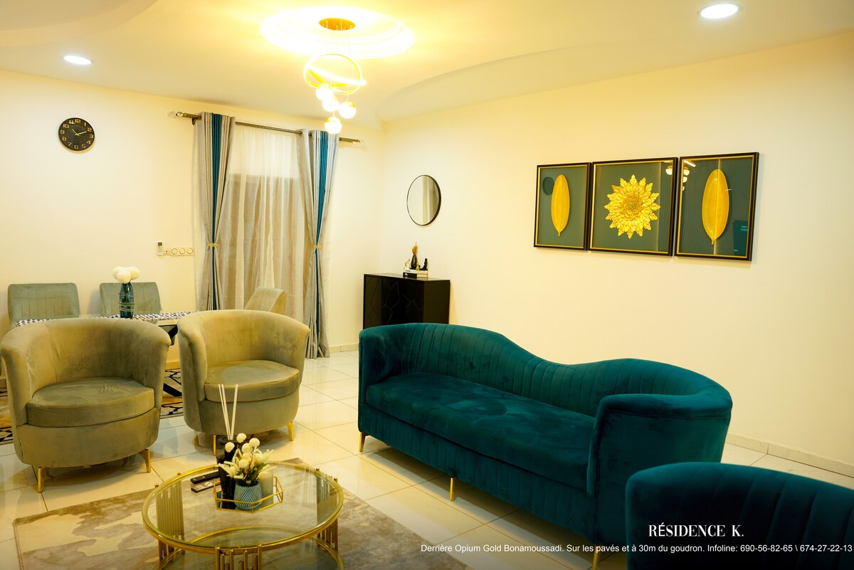 A comfortable living area is highlighted by a plush turquoise sofa and modern accent chairs. A glass coffee table is positioned in front of a decorative wall featuring two floral art pieces. Natural light streams in through large windows, complemented by elegant curtains.