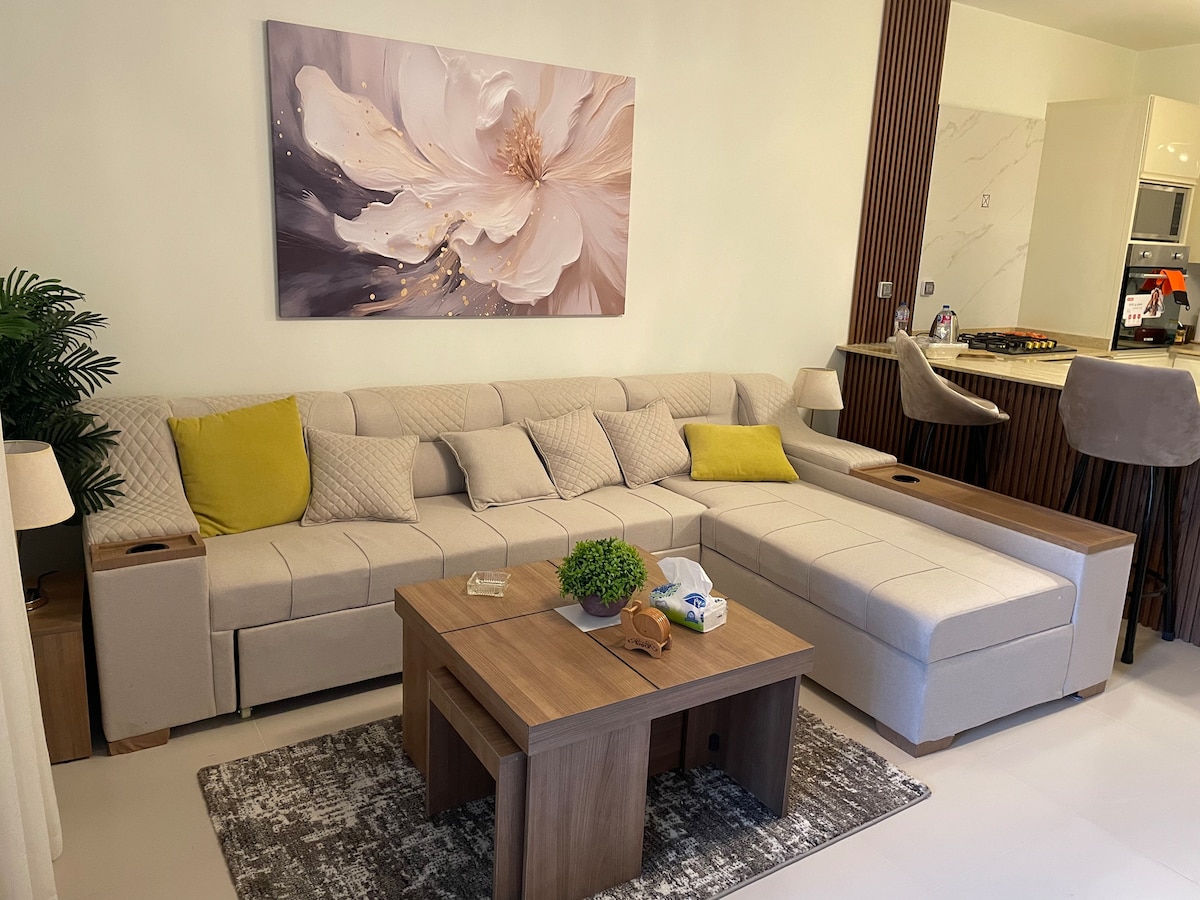 A spacious living area is furnished with a light-colored sectional sofa, complemented by decorative cushions. A wooden coffee table sits atop a patterned area rug, while a potted plant and soft lighting fixtures add a serene touch. In the background, a sleek kitchen is partially visible.