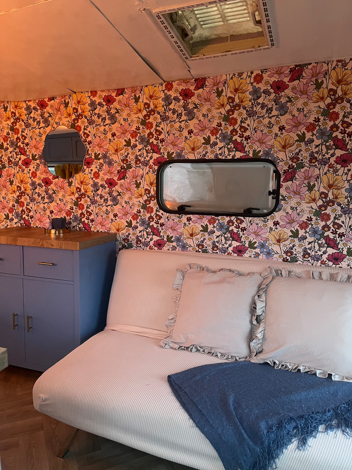 A well-decorated interior features floral wallpaper in vibrant colors. A light-colored couch adorned with decorative throw pillows offers comfortable seating. A small, matching blue cabinet is positioned nearby, while a round mirror reflects light from the ceiling vent above.