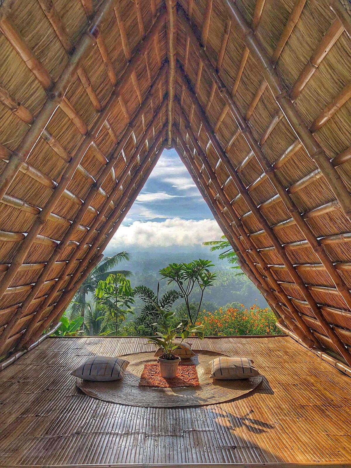 A triangular-shaped interior features vaulted bamboo ceilings and a panoramic view of the surrounding greenery and clouds. A round floor mat with a potted plant and decorative items is centered on the wooden floor, complemented by two plush floor cushions.