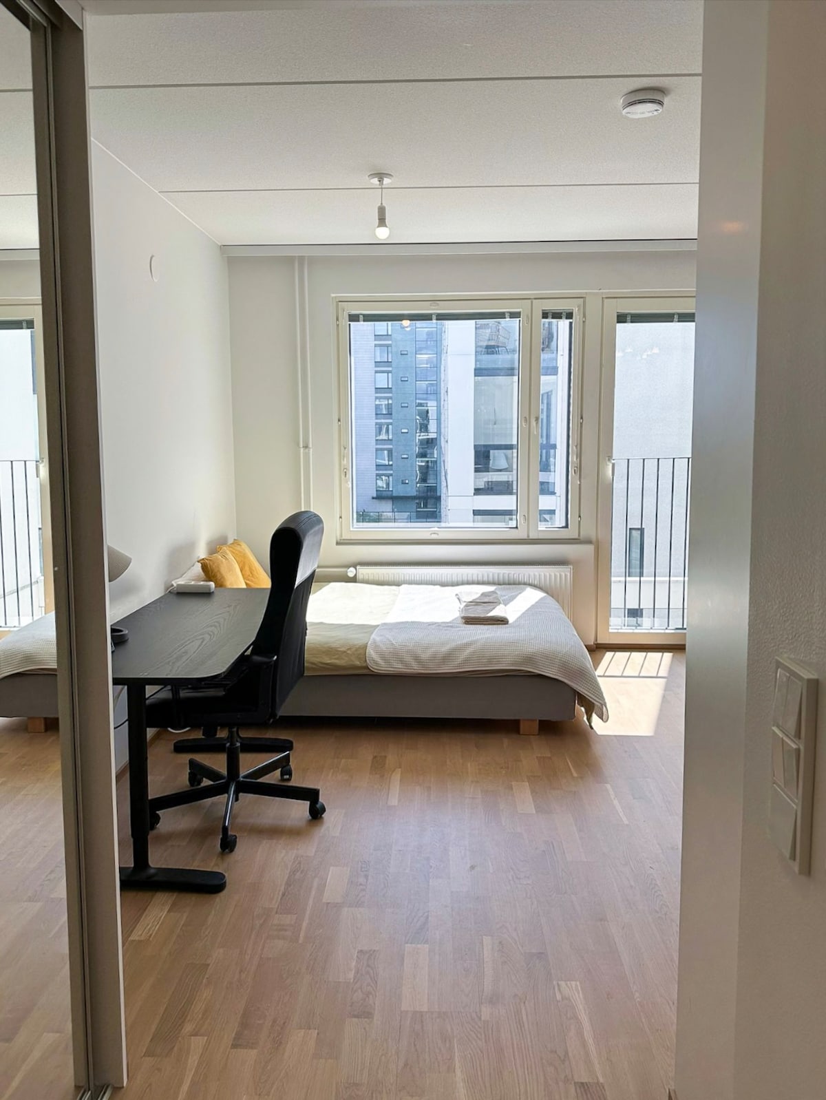 A bright room is visible with a spacious bed and a desk chair positioned at a small desk. Large windows allow natural light to fill the space, with a view of the neighboring buildings. A soft yellow cushion and a book are placed on the bed.