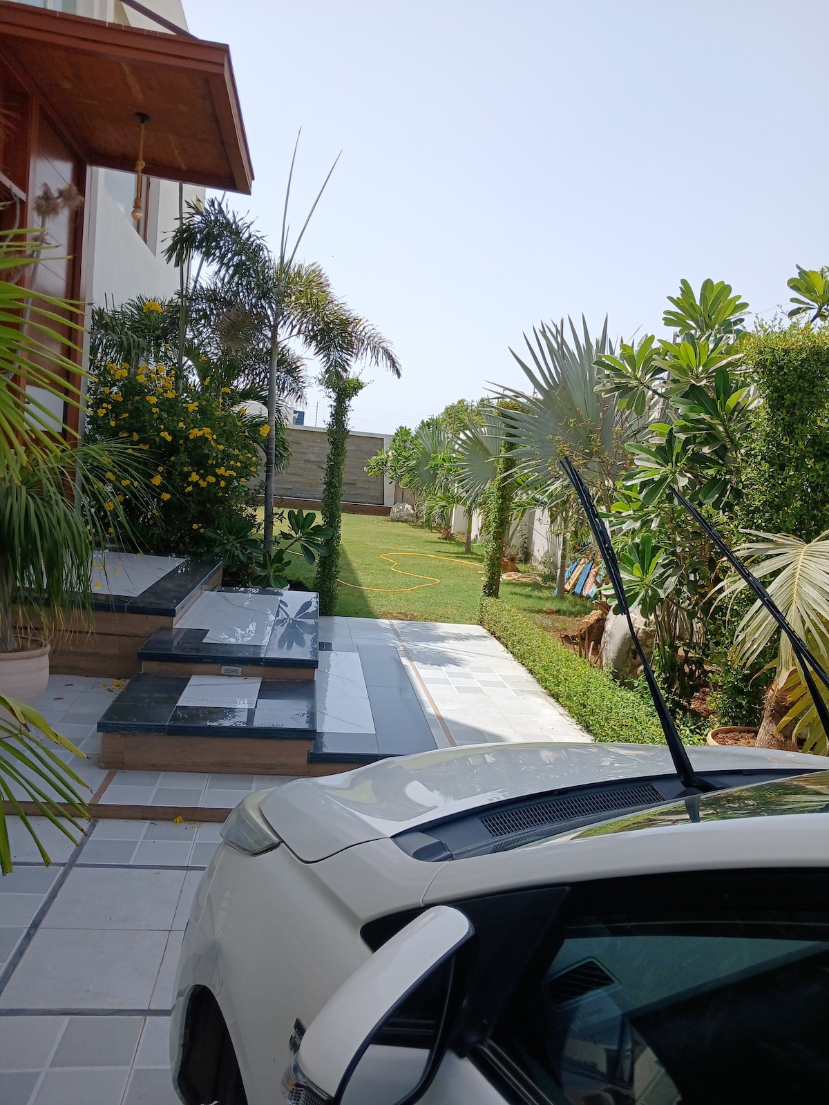 A well-maintained pathway leads towards a lush garden, framed by vibrant greenery and palm trees. The driveway features a car parked on a tiled surface, with steps leading up to the entrance of the property. The bright sky enhances the lively atmosphere.