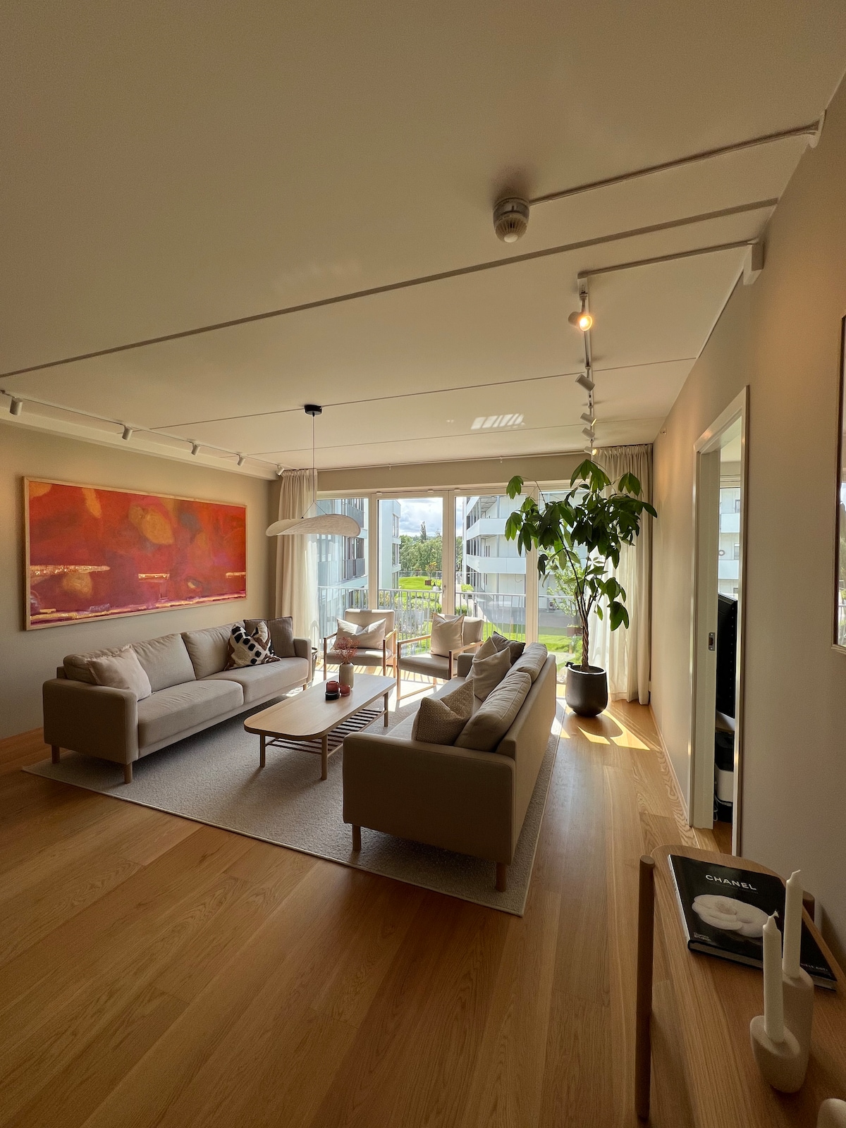A bright living room features large windows that allow natural light to fill the space. A neutral-toned sofa is centered on a light area rug, accompanied by a coffee table. A tall plant adds a touch of greenery, and a piece of abstract art decorates the wall.