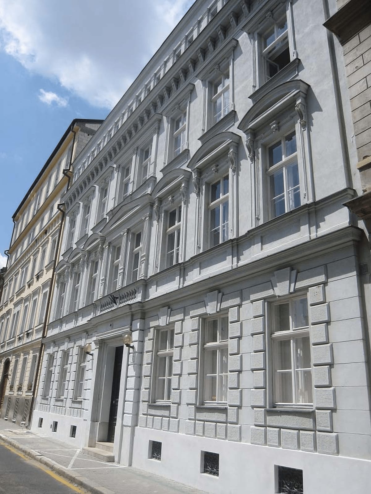 The exterior of a historic building is presented, showcasing a facade characterized by ornate detailing and large windows. Light reflects off the stone surface under a bright blue sky, highlighting the architectural features of the structure.