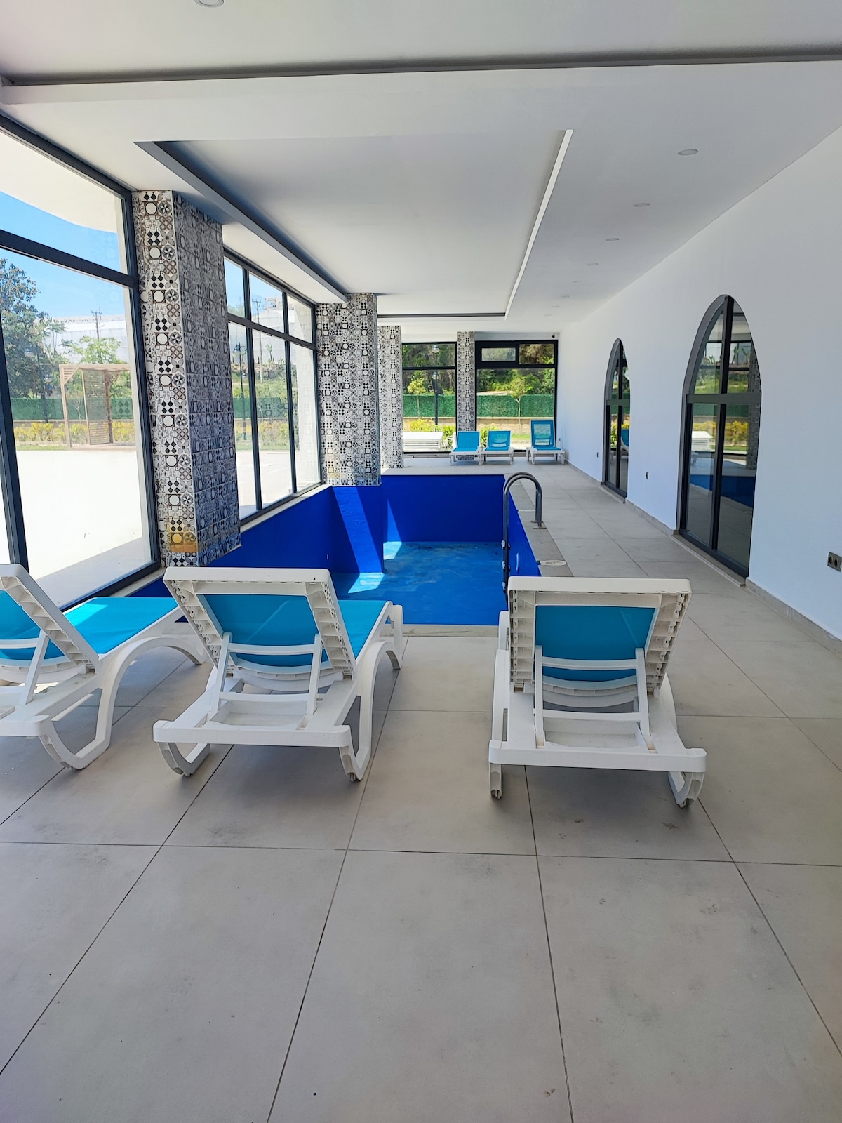 An indoor area features a rectangular blue pool surrounded by large glass windows. Two lounge chairs with bright blue cushions are positioned nearby, offering comfort for relaxation. Natural light fills the space, highlighting the contemporary design and the tiled accent wall.