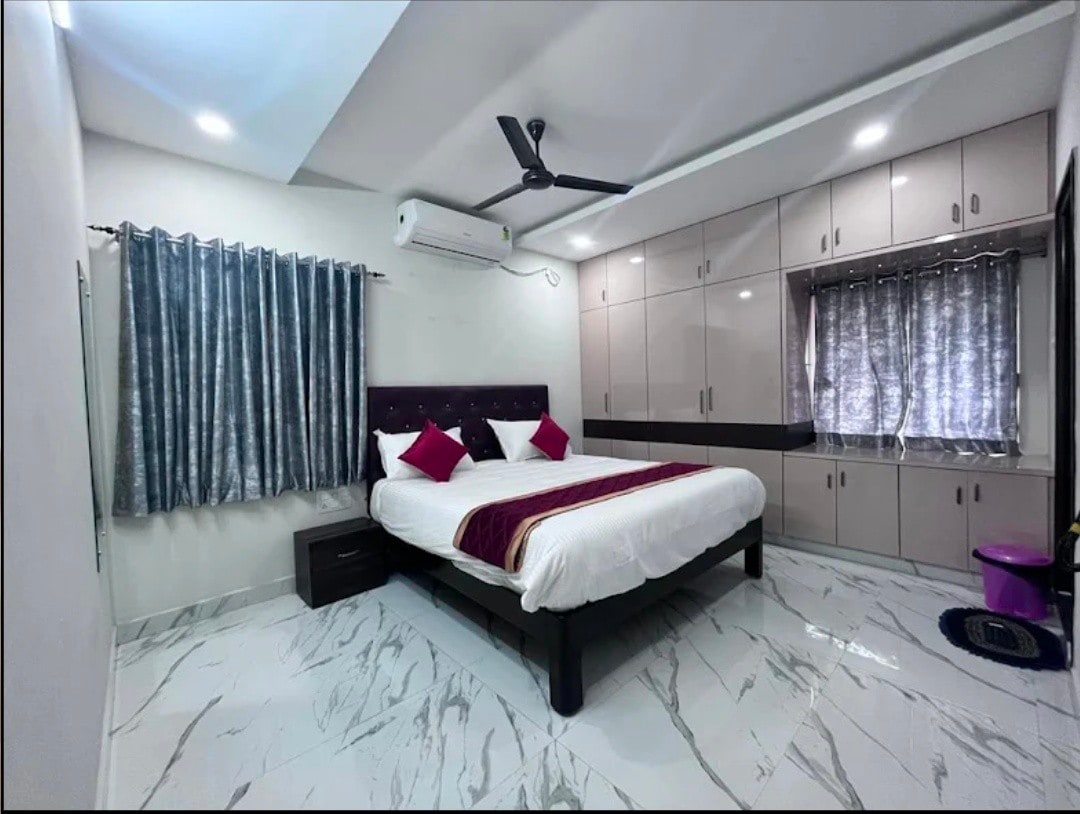 A spacious bedroom features a large bed with white linens and accent pillows in shades of red. A fan is mounted on the ceiling, and natural light filters through partially draped windows. Sleek cabinetry lines one wall, providing ample storage space.