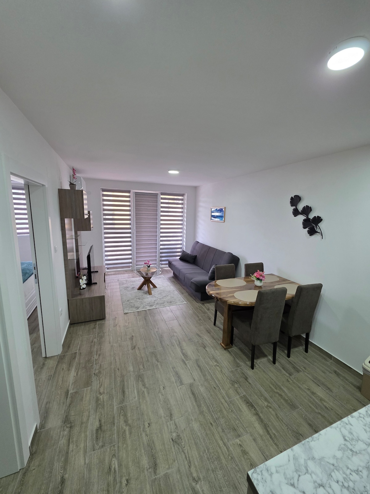 A modern living space is presented with a comfortable gray sofa and a round wooden coffee table atop a textured rug. A dining area with a two-person table and chairs is included. Natural light enters through the slatted window coverings, enhancing the inviting atmosphere.