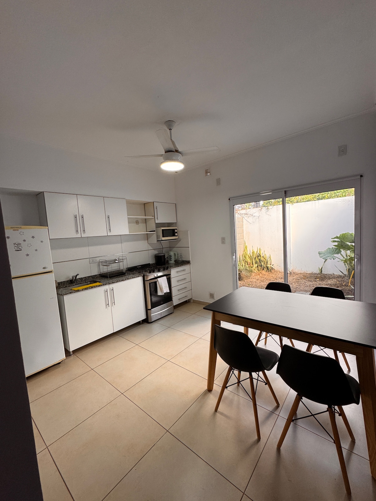 A modern kitchen is equipped with essential appliances, including a refrigerator, microwave, and oven. A dining table with four black chairs occupies the space. Natural light filters through a sliding glass door, revealing a small outdoor area bordered by plants.