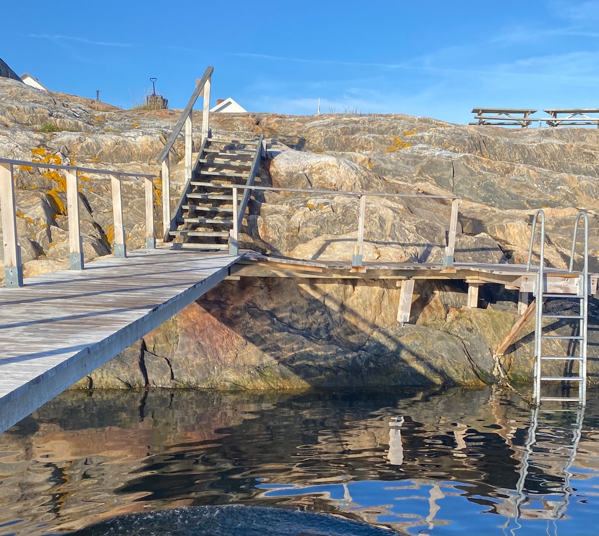 A wooden staircase leads up from a rocky shore to a higher area, flanked by smooth stones. A ladder provides access to the water, where reflections of the clear sky and surrounding nature can be seen in the calm surface.
