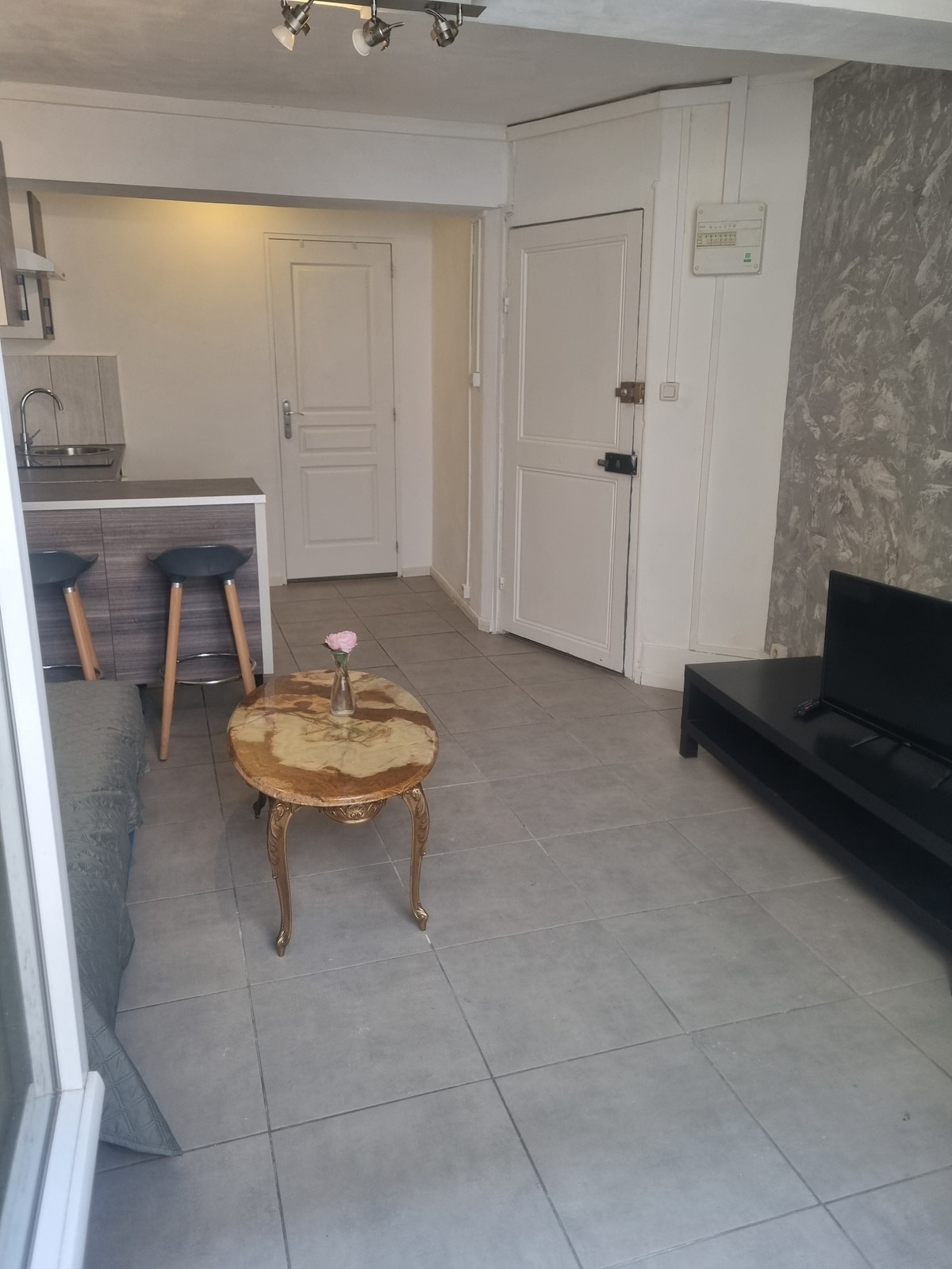 A compact living space is arranged with a small wooden table and two bar stools beside a kitchenette. A grey couch is positioned next to a modern black television on a low stand. Light-colored tiles cover the floor, enhancing the room's spacious feel.