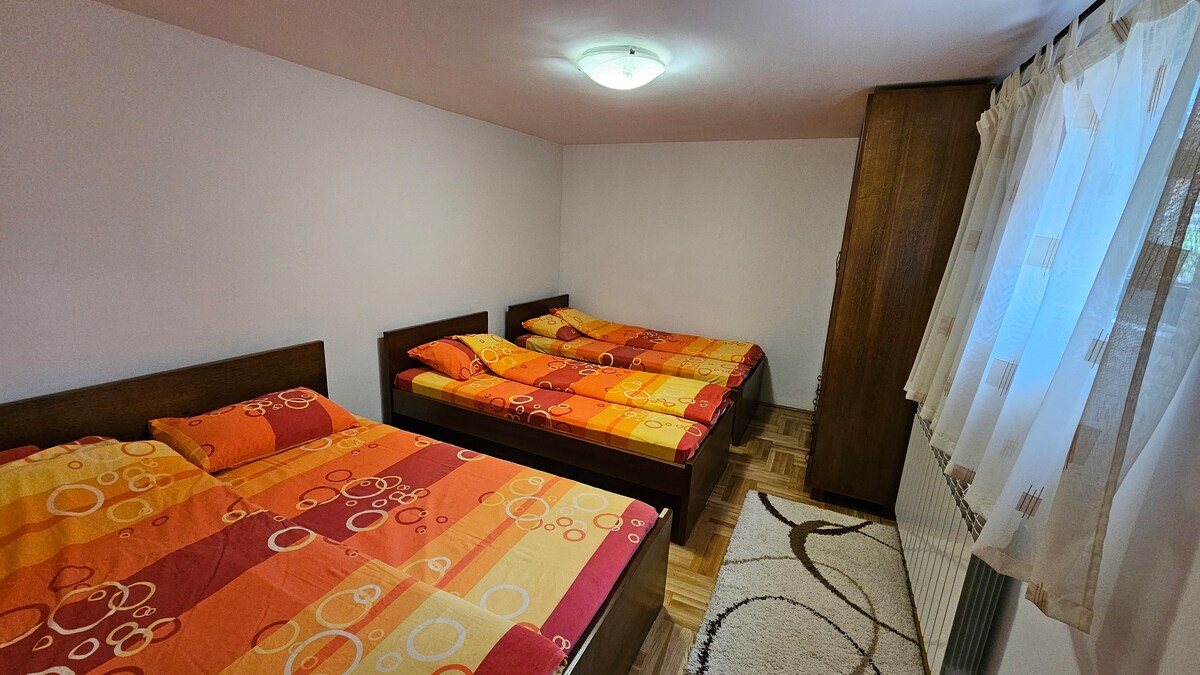 A sleeping area is observed with three spacious beds featuring bright orange and yellow bedding. A patterned rug is placed on the wooden floor, and a wardrobe is positioned against one wall. Natural light enters through a window adorned with light curtains.