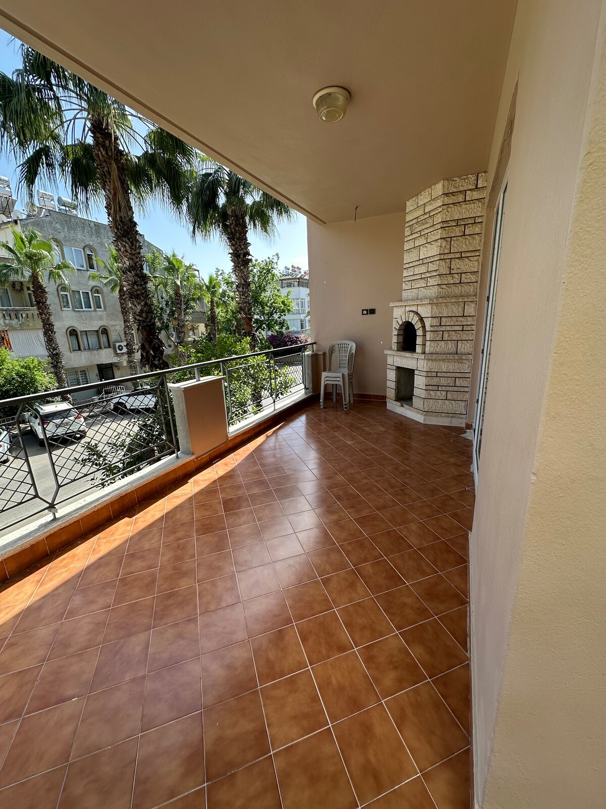 A spacious balcony is shown, featuring tiled flooring and a built-in stone barbecue. Lush palm trees can be seen outside, providing a green backdrop. The railing offers views of the surrounding area, creating an open and airy atmosphere.