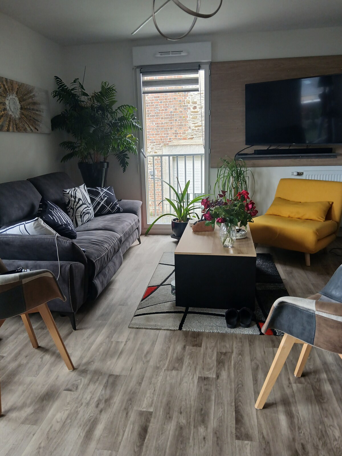 A living area features a dark gray sofa adorned with patterned cushions. A bright yellow armchair sits next to a low coffee table. Green plants are placed beside large windows, allowing natural light into the space, complemented by a decorative area rug and a mounted television.