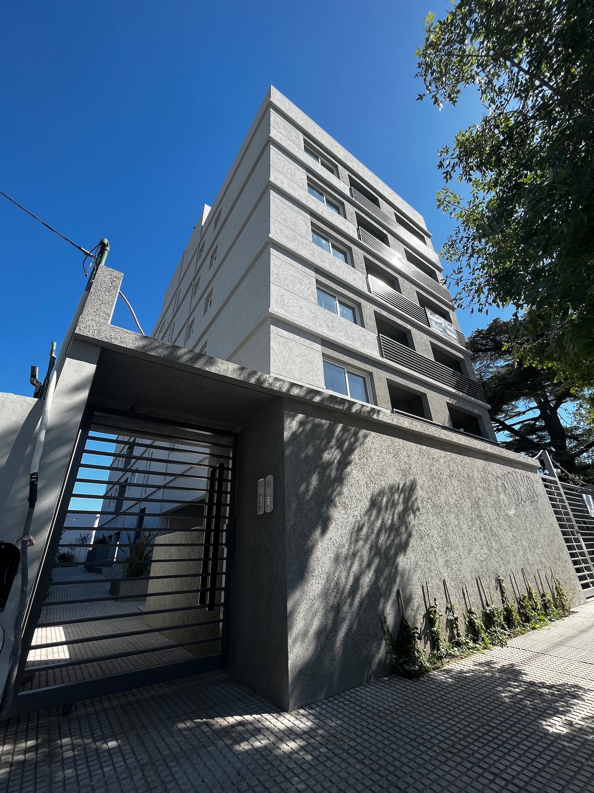 The exterior of a modern, multi-story building is shown, featuring a sleek, minimalist design. A secure entrance is visible, framed by a stone wall. Clear blue skies contrast with the building's neutral tones, and lush greenery is seen along the perimeter.