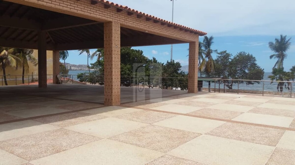 An open outdoor area is visible, featuring a spacious tiled surface shaded by a roof supported by sturdy columns. Tropical palm trees and ocean views create a serene backdrop, with the gentle movement of grass and foliage suggestive of a coastal environment.