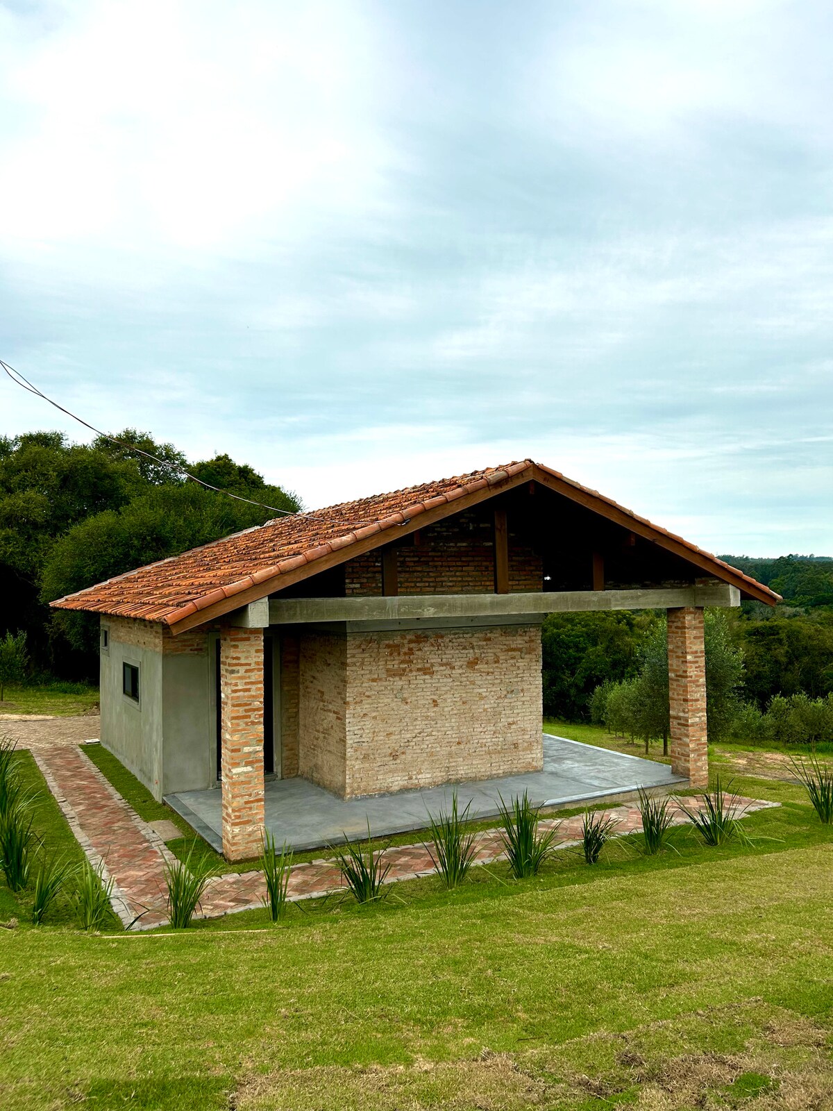 A rustic house features a sloped roof with terracotta tiles and a brick facade. The open area under the roof offers shaded space, while the surrounding landscape includes green grass and gentle foliage. The structure is positioned amidst a tranquil rural setting.