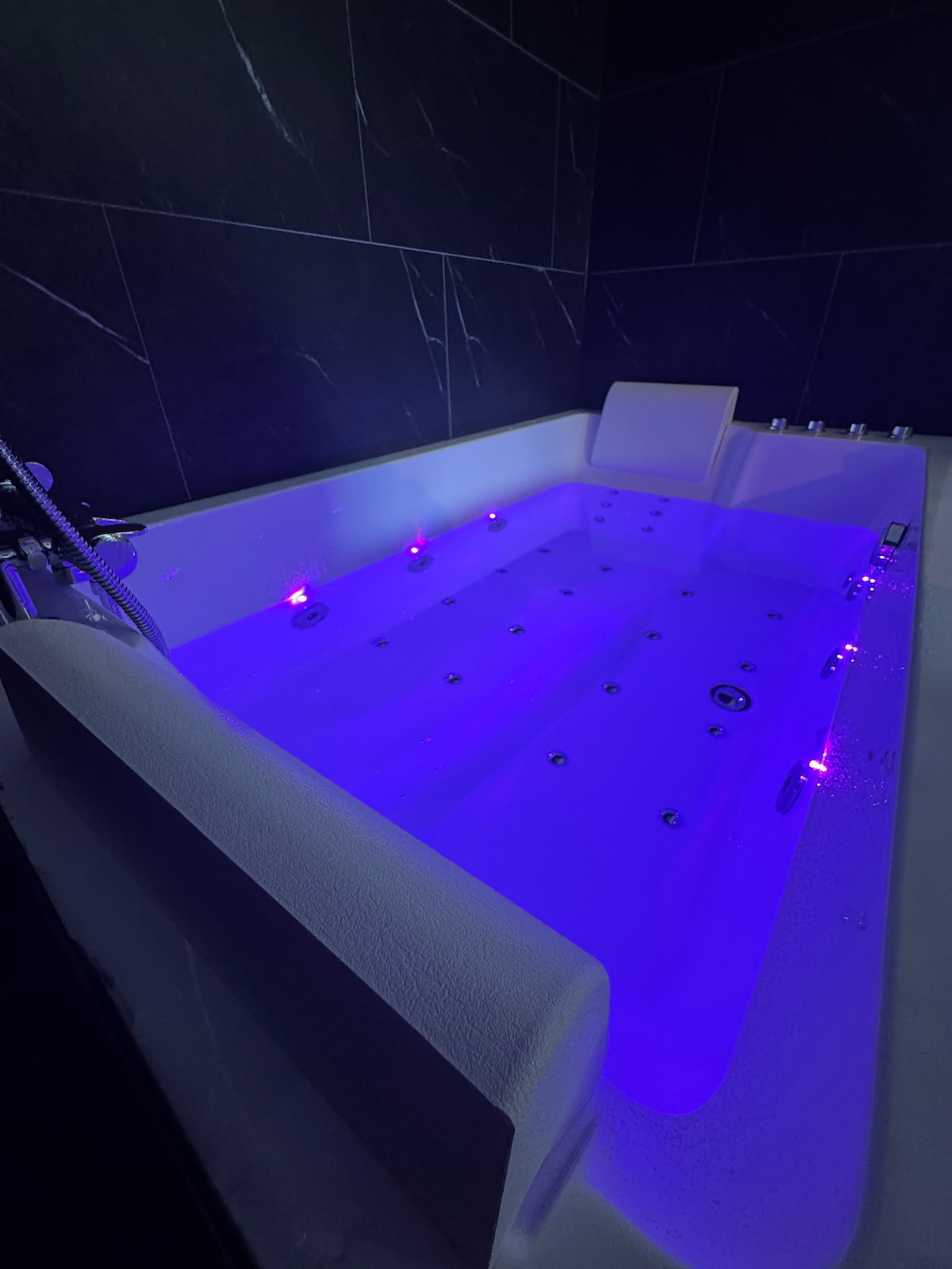A white, two-person whirlpool bathtub is depicted, featuring illuminated jets in soothing purple tones. The surrounding area is lined with dark, textured tiles, creating a modern and intimate atmosphere. The sleek design invites relaxation and tranquility.