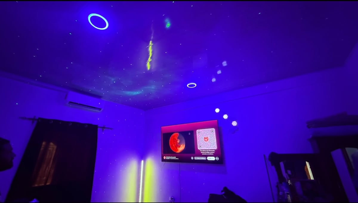 The ceiling is adorned with a starry projection, creating a celestial ambiance. A television screen displays content in the foreground, while soft lighting complements the room's modern decor. An air conditioning unit is visible, ensuring a comfortable environment.