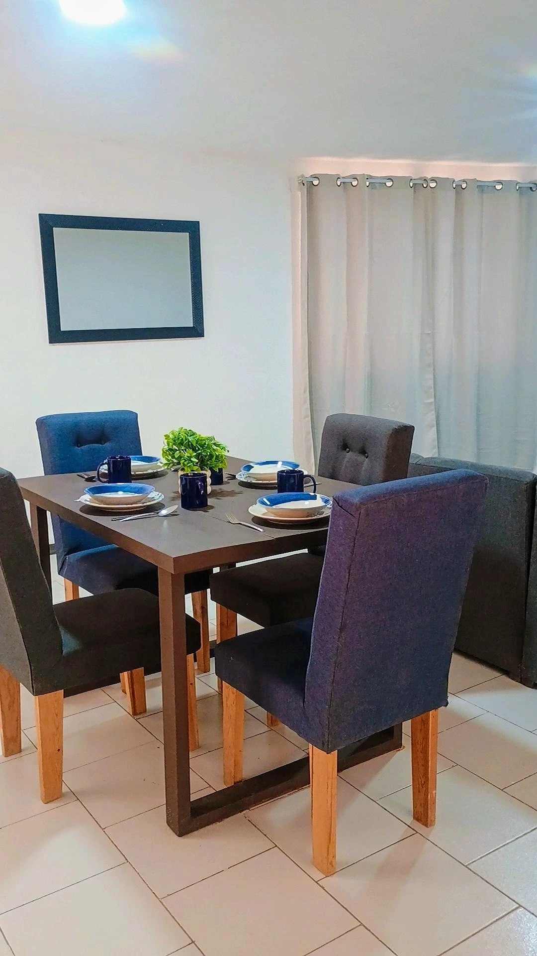 A dining area is presented with a wooden table surrounded by four high-backed chairs in dark fabric. The table is set with blue dishware and a small greenery centerpiece. A mirror hangs on the wall, reflecting the space, with light filtering in through sheer curtains.