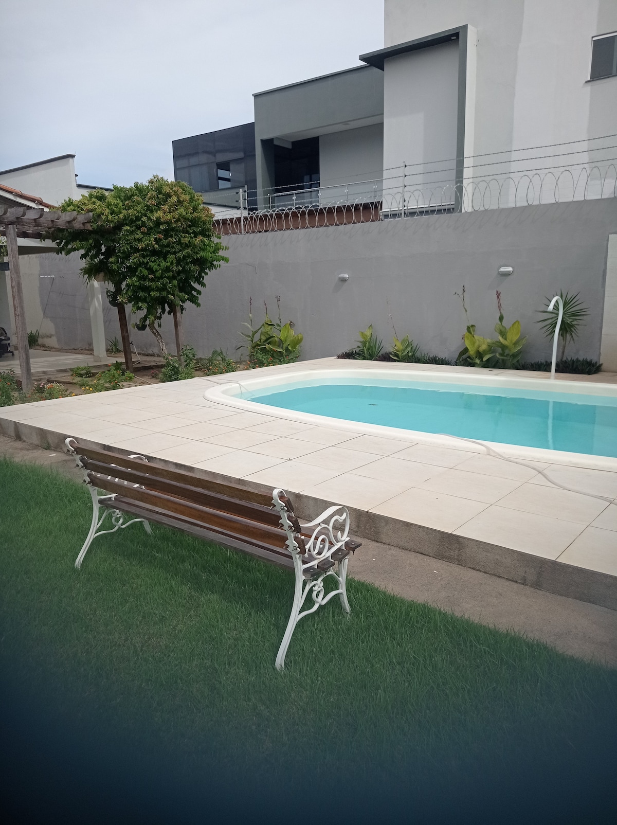A serene outdoor space features a clear swimming pool surrounded by neatly maintained greenery. A bench is positioned on a patio made of light-colored tiles, offering a comfortable spot to relax while enjoying the view of the pool and landscaped garden.