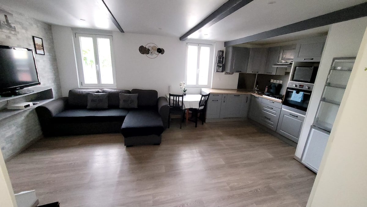 A modern living space features a comfortable dark sofa facing a wall-mounted television. A dining table with chairs is visible near the kitchen, which includes sleek cabinetry and built-in appliances. Natural light enters through two windows, complementing the light wood flooring.
