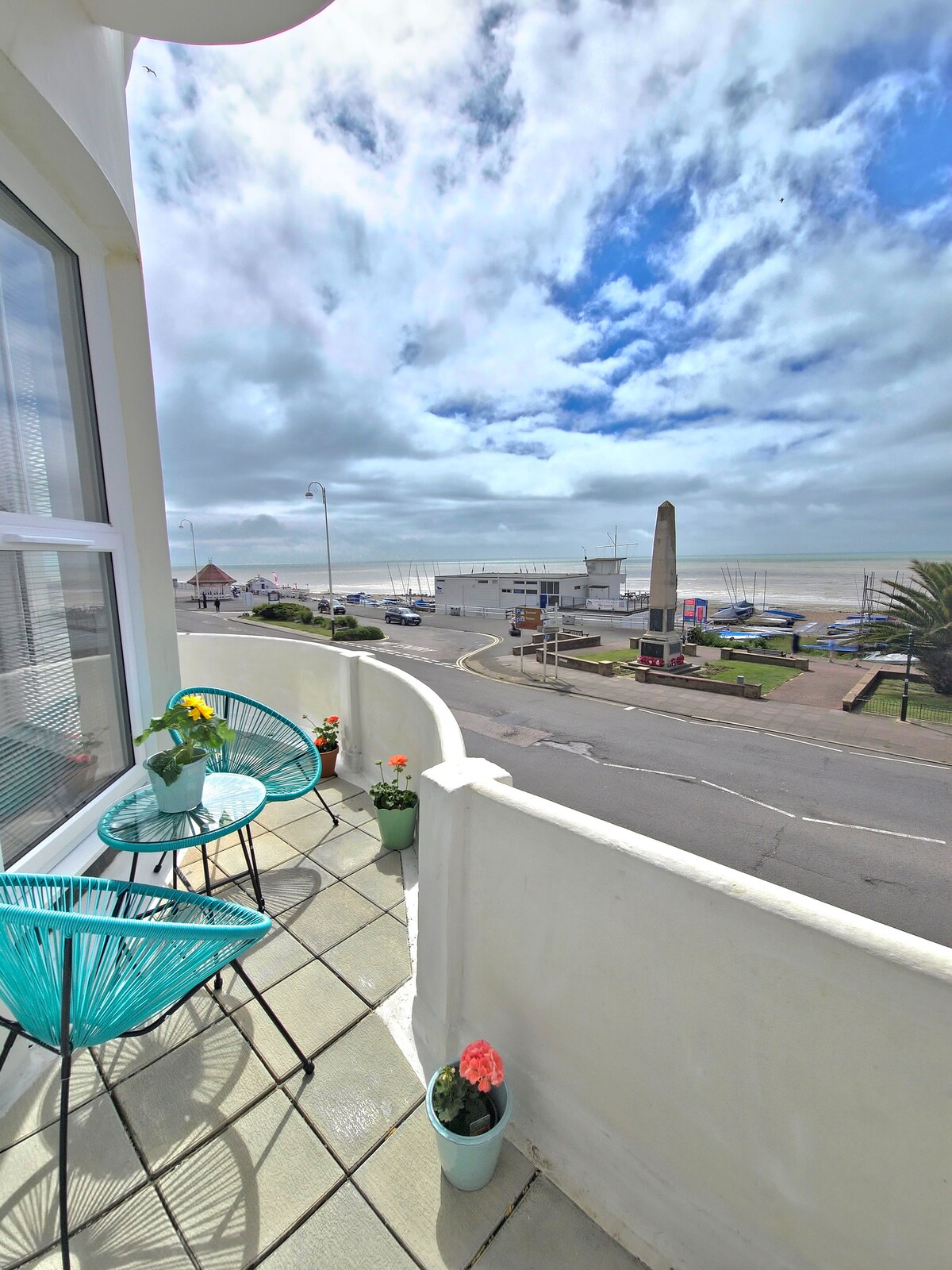 A balcony space features two teal chairs beside small potted flowers. A view of the coast is visible, with the sea and distant boats framed by a cloudy sky. Sidewalk and street activity can be observed, enhancing the vibrant outdoor atmosphere.