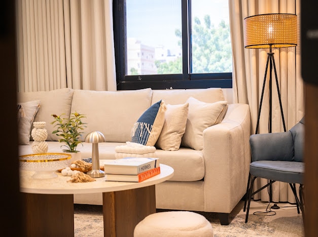 A light-filled sitting area features a spacious beige couch adorned with decorative pillows. A round coffee table is complemented by decorative items and a small green plant. A sleek floor lamp stands beside a blue accent chair, enhancing the cozy atmosphere.