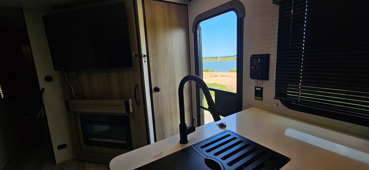 An open kitchen area is visible, featuring a modern sink and countertop. A television is mounted on a wooden wall unit, and a window frames a view of the water and landscape outside. Natural light filters through the window's dark blinds.