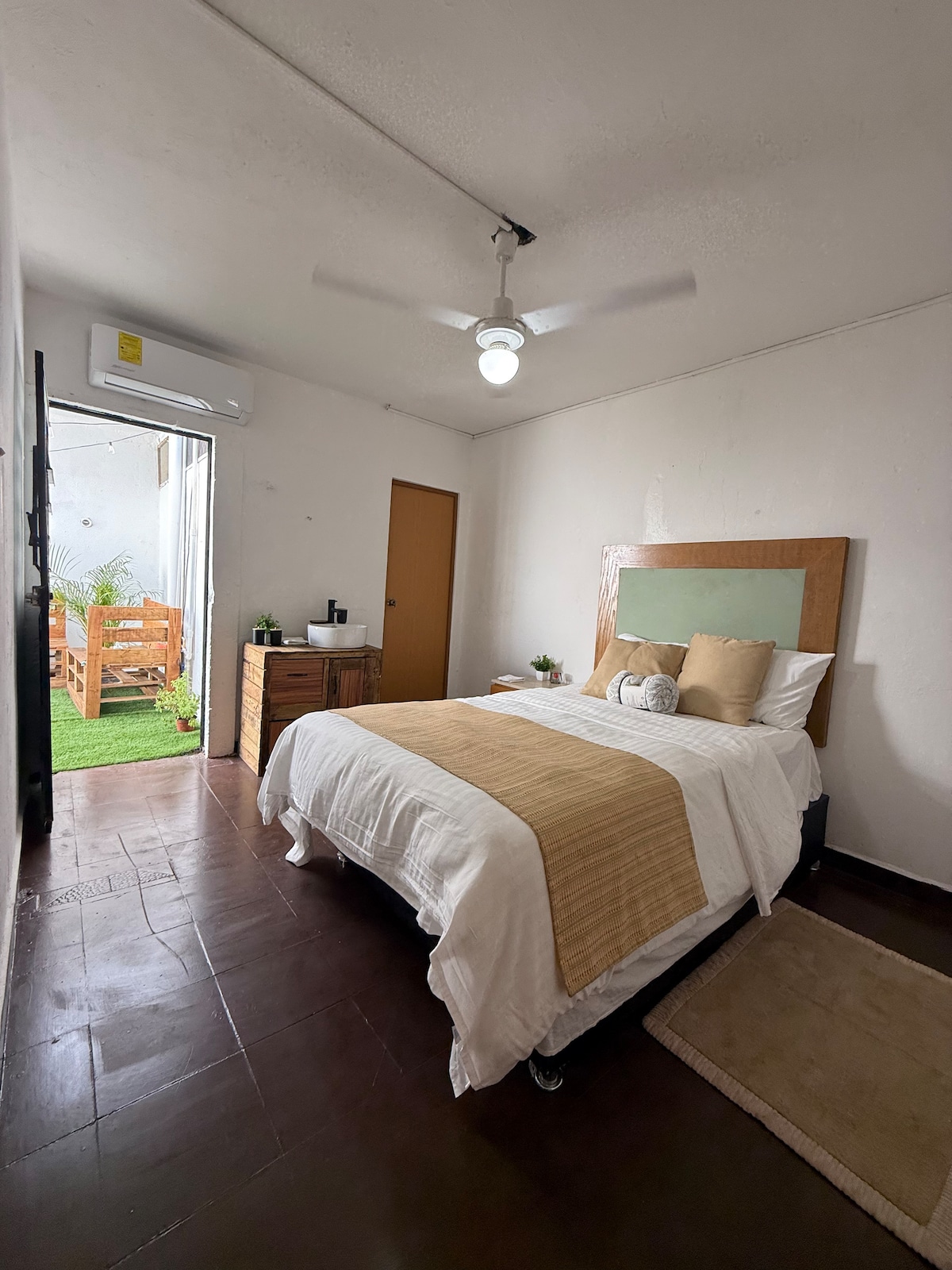 A cozy bedroom features a spacious bed with neutral-toned bedding and decorative pillows. A ceiling fan provides ventilation, and light enters through a nearby window. Across the room, a wooden nightstand holds small plants, while a doorway leads to an outdoor area with seating.