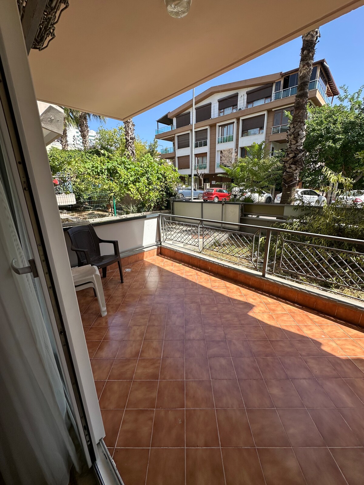 A bright balcony area is visible, featuring tiled flooring and a few potted plants. A single chair is positioned near the railing, providing an outdoor space with views of neighboring greenery and buildings under a clear blue sky.