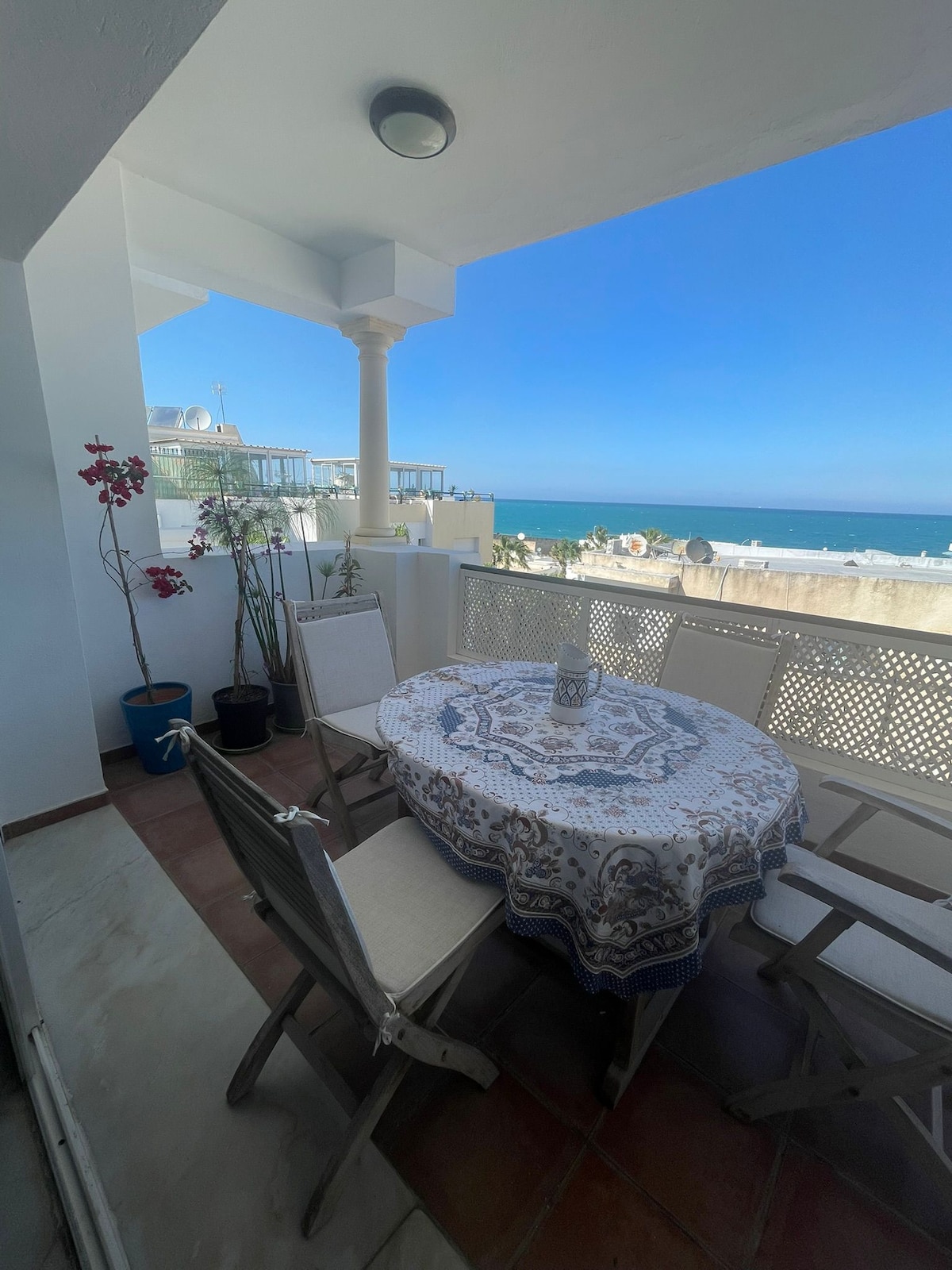 An inviting terrace features a round table covered with a decorative tablecloth and surrounded by wooden chairs. Potted plants add a touch of greenery, while the expansive view of the sea is visible in the background under a clear blue sky.