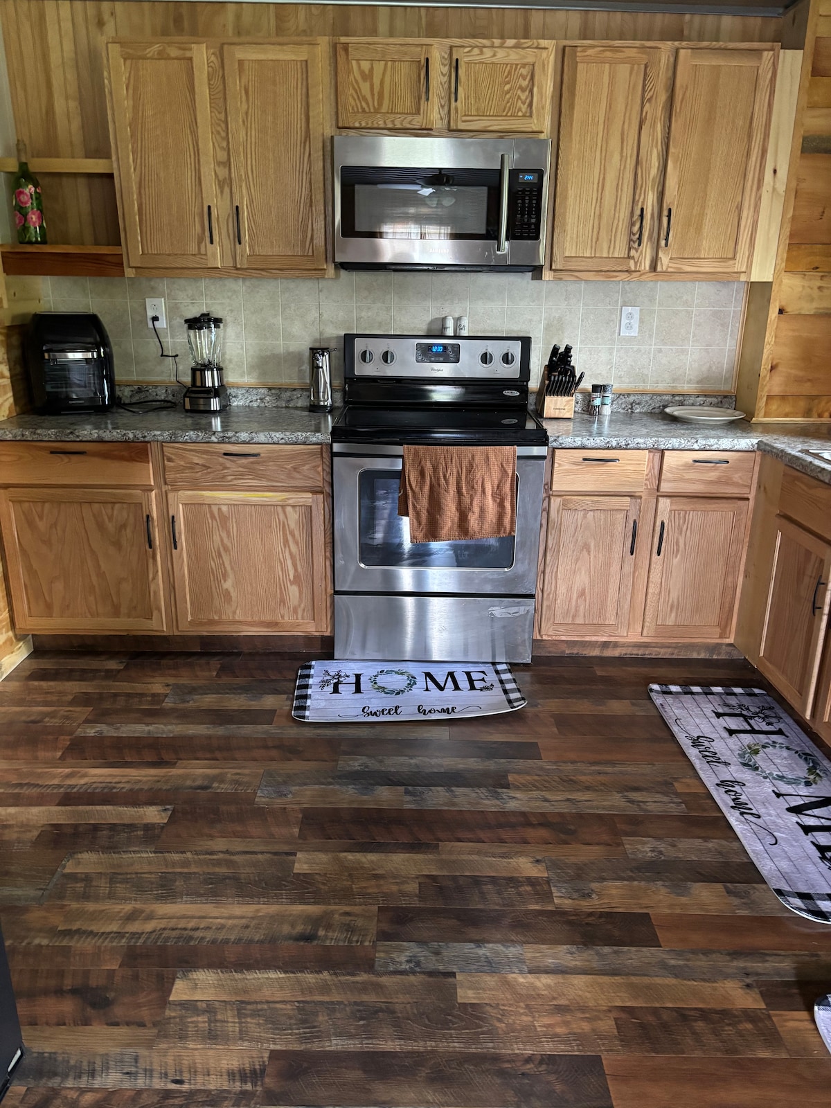 The kitchen features wooden cabinetry and sleek countertops, equipped with modern stainless steel appliances including a refrigerator and oven. Decorative rugs add a homely touch to the floor, while ample lighting highlights the workspace. A coffee maker and blender are conveniently placed on the counter.