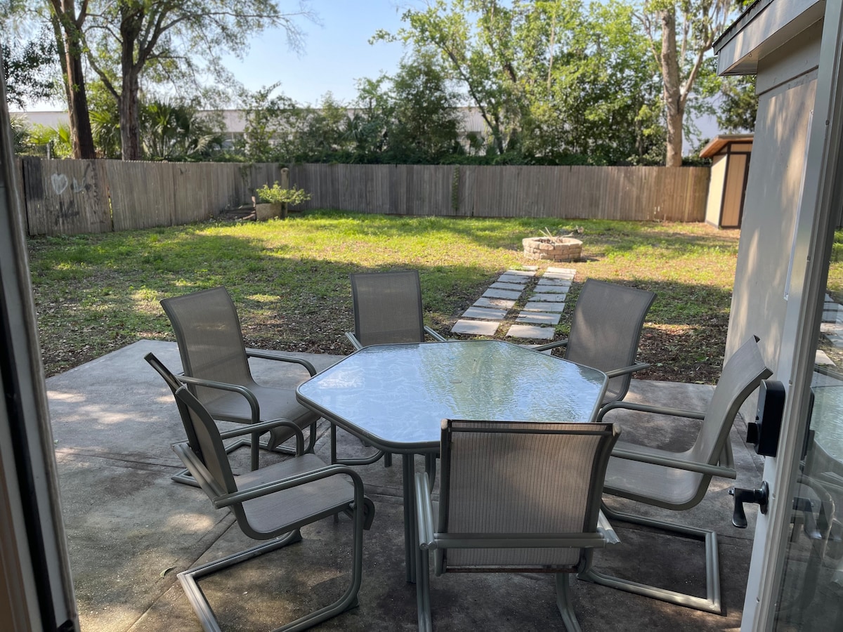 An outdoor patio area is depicted, featuring a round table surrounded by six metal chairs on a concrete surface. The lush green yard extends beyond, adorned with scattered stepping stones and bordered by a wooden fence. Sunlight filters through the trees, creating a bright and open setting.
