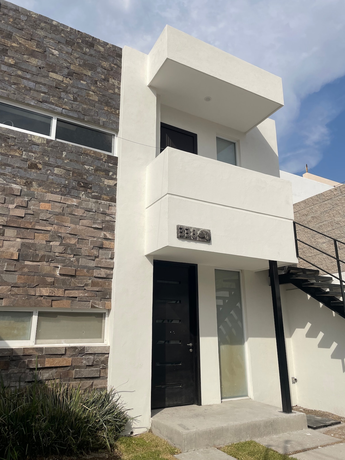 The entrance of a contemporary loft is shown, highlighting a clean white façade and stone accents. A dark door provides access, and a staircase leads to the upper level. Large windows enhance natural light, while greenery adds a touch of nature to the entrance.
