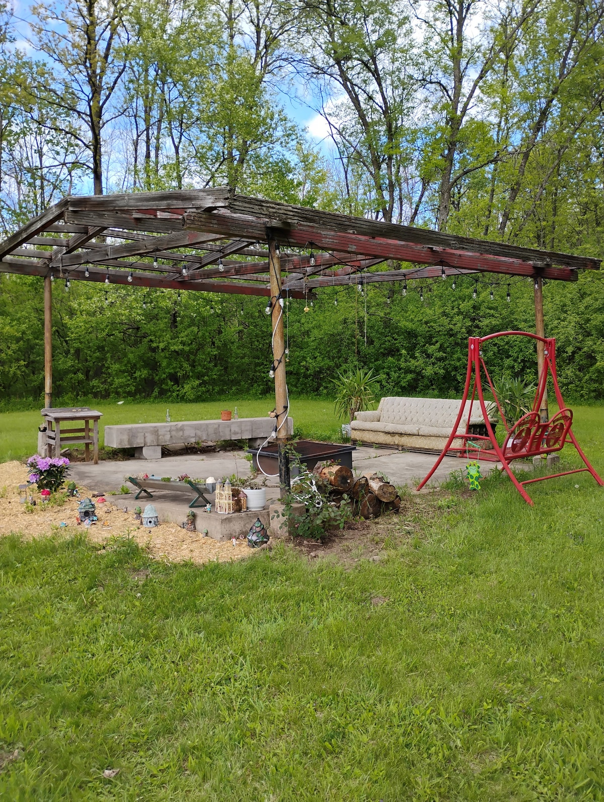 An outdoor seating area features a covered frame with string lights overhead. A stone fire pit is surrounded by a circular seating arrangement, complemented by a red swing chair and a sofa. Lush greenery and decorative items add touches of nature.