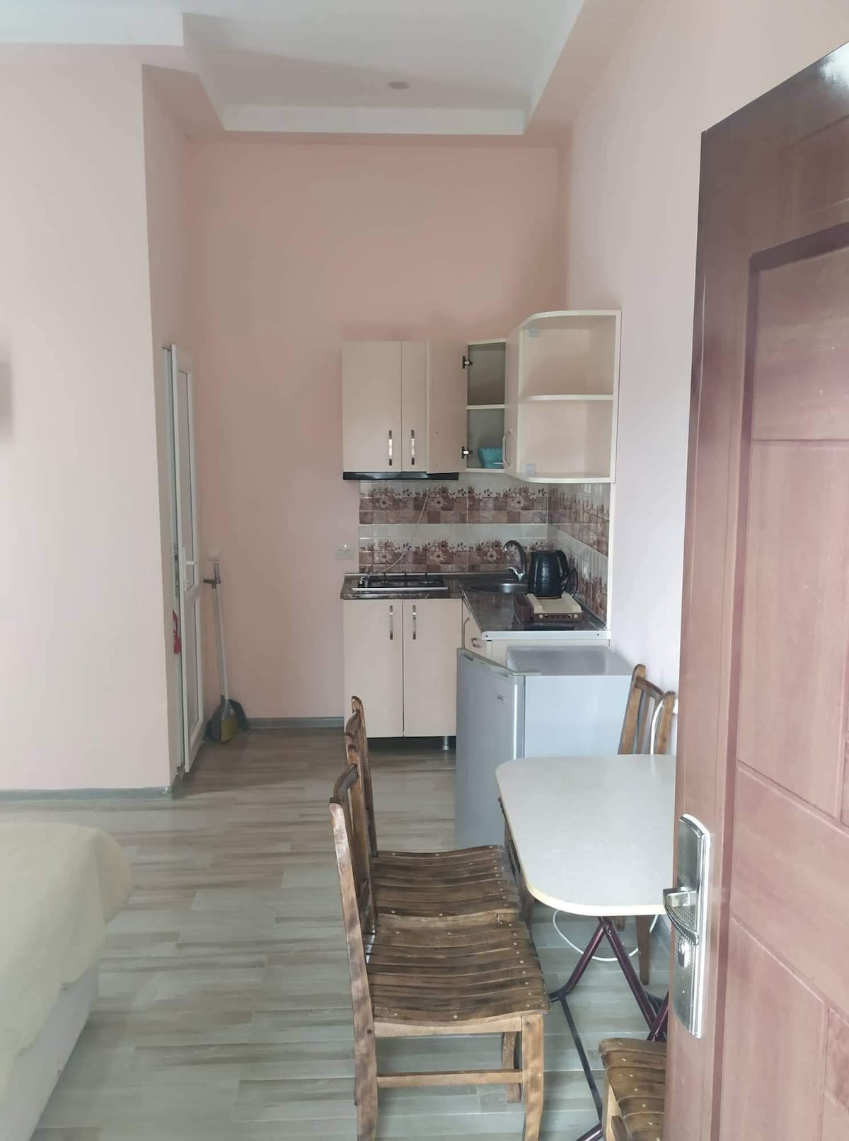 A compact kitchen area is visible, featuring light-colored cabinets and essential appliances, including a stove and refrigerator. A small dining table with two wooden chairs is positioned nearby, contributing to the efficient use of space. Soft pink walls provide a gentle backdrop.