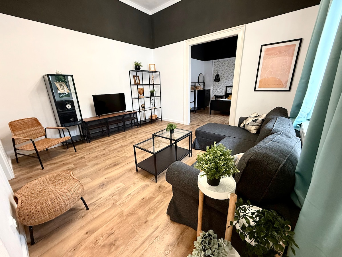 A modern living area features a comfortable gray sofa and a coffee table, with light wooden flooring enhancing the spacious feel. A black shelving unit holds decorative items and a television is positioned against the wall. Natural light enters through large windows adorned with curtains.