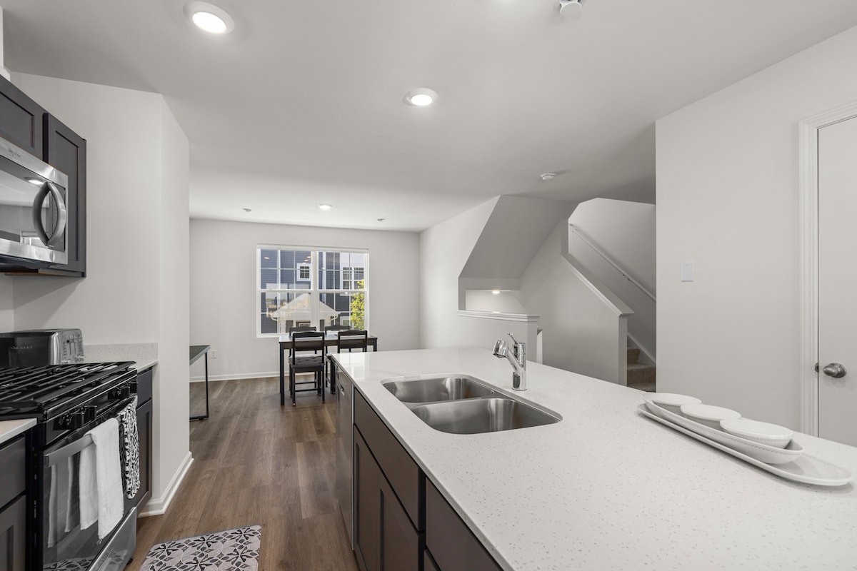 A modern kitchen is featured with dark cabinetry and stainless steel appliances, including a gas stove and built-in microwave. A spacious countertop with a double sink and stylish tableware is visible. Natural light fills the open area, connecting to the dining space and staircase beyond.
