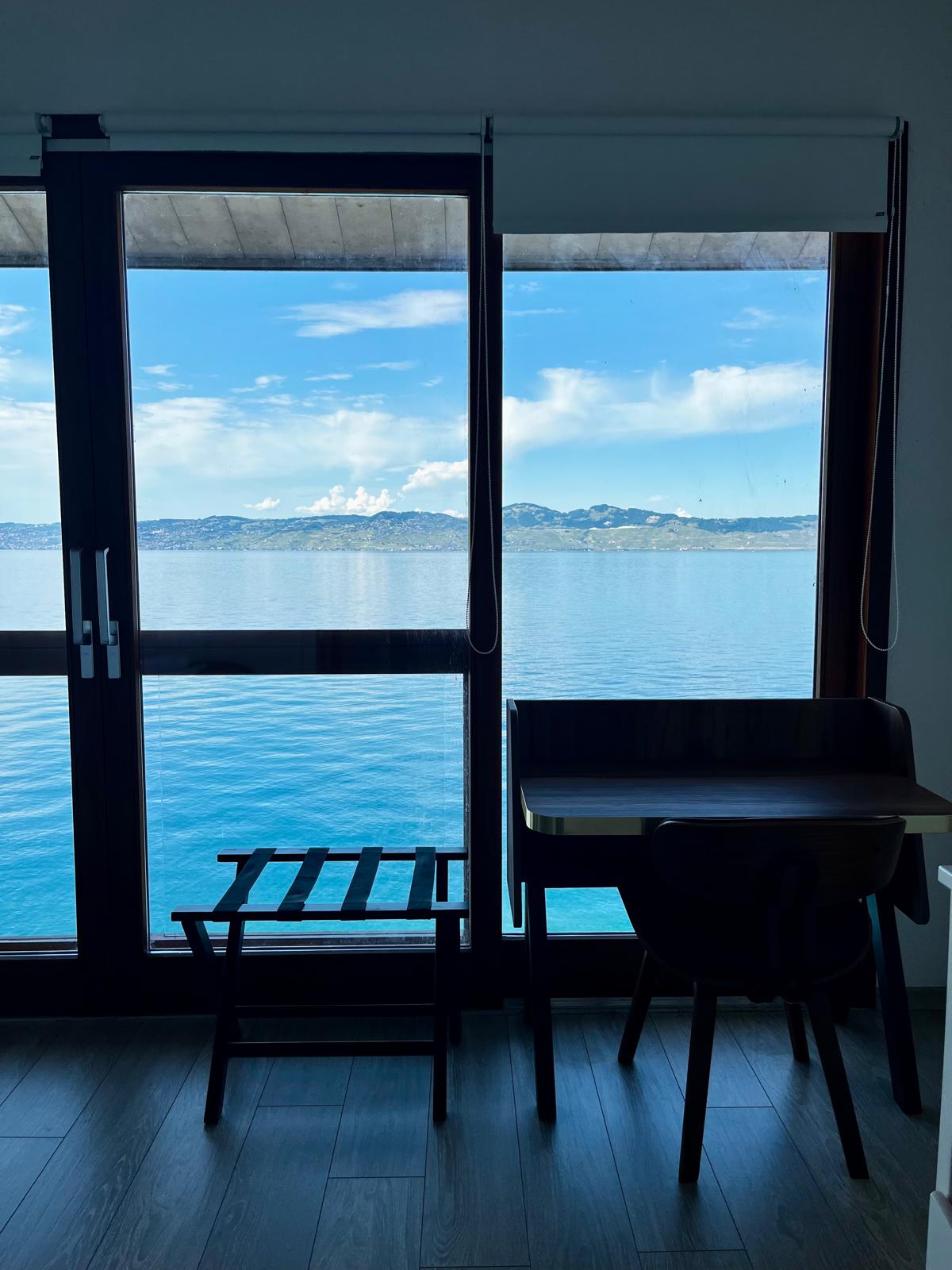 A serene view is captured through large glass doors overlooking the calm waters of Lake Geneva. A small desk and a chair accompany a wooden lounger, creating a quiet space for reflection. The distant Swiss Alps can be seen gently rising on the horizon.