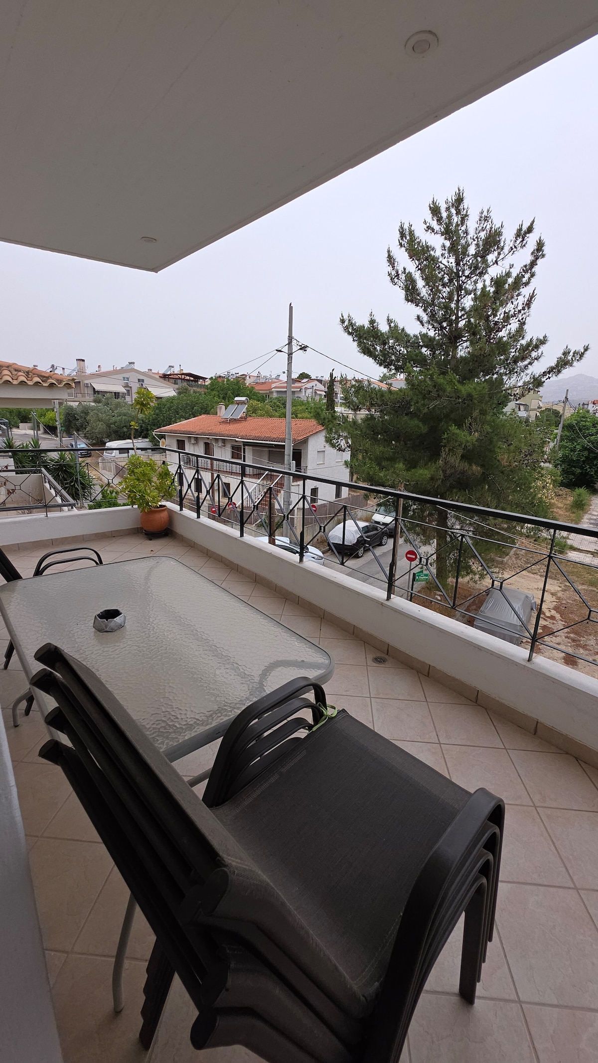 A spacious balcony features a glass dining table surrounded by five chairs. A potted plant adds a touch of greenery, and a partial view of surrounding trees and buildings is visible. The area is designed for outdoor relaxation, providing a serene environment.