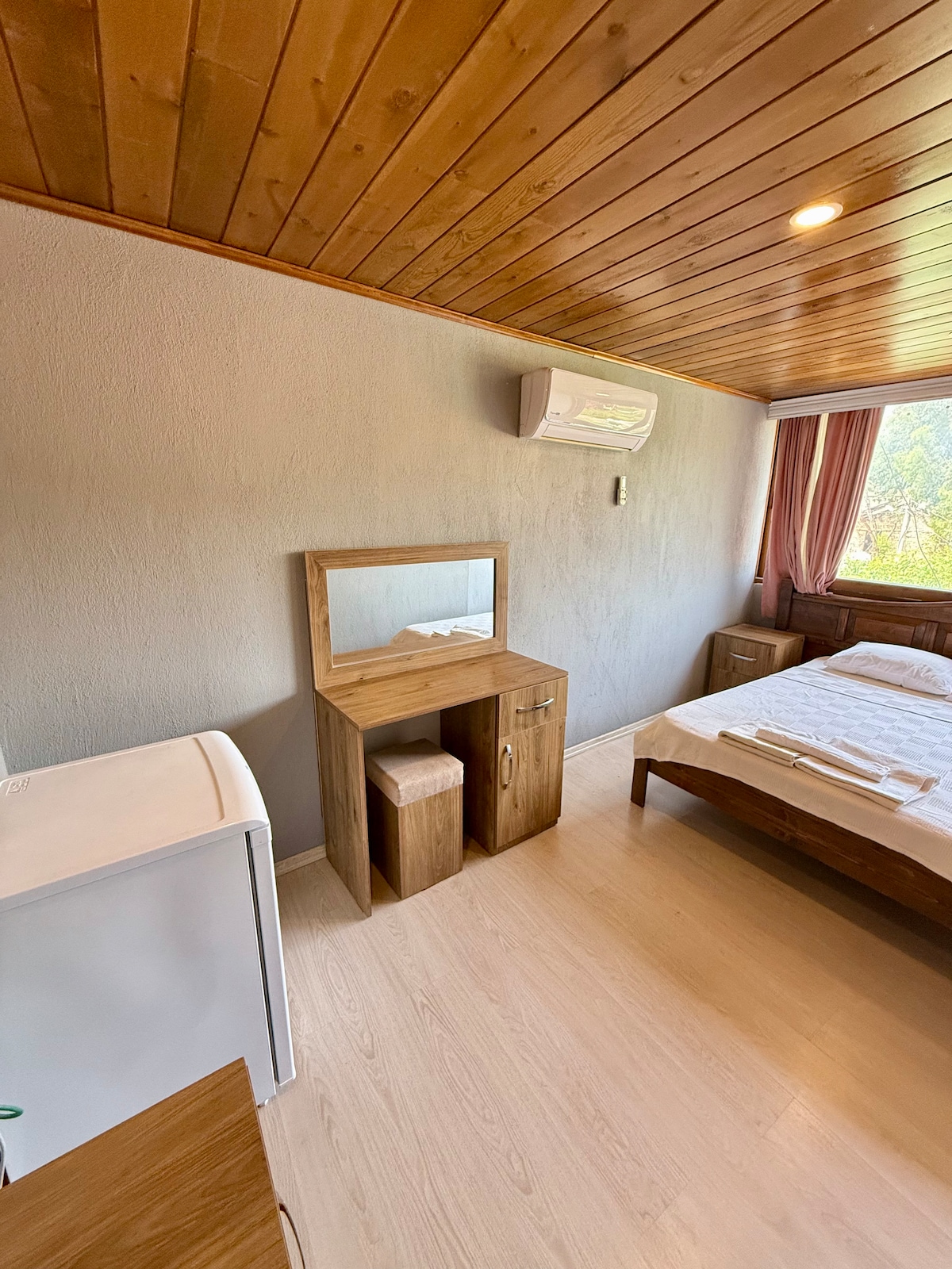 The room features clean, minimalistic design elements including a spacious bed with fresh linens and bedside tables. A compact refrigerator and a wooden vanity with a mirror are present. Natural light enters through the window, complemented by soft curtains.