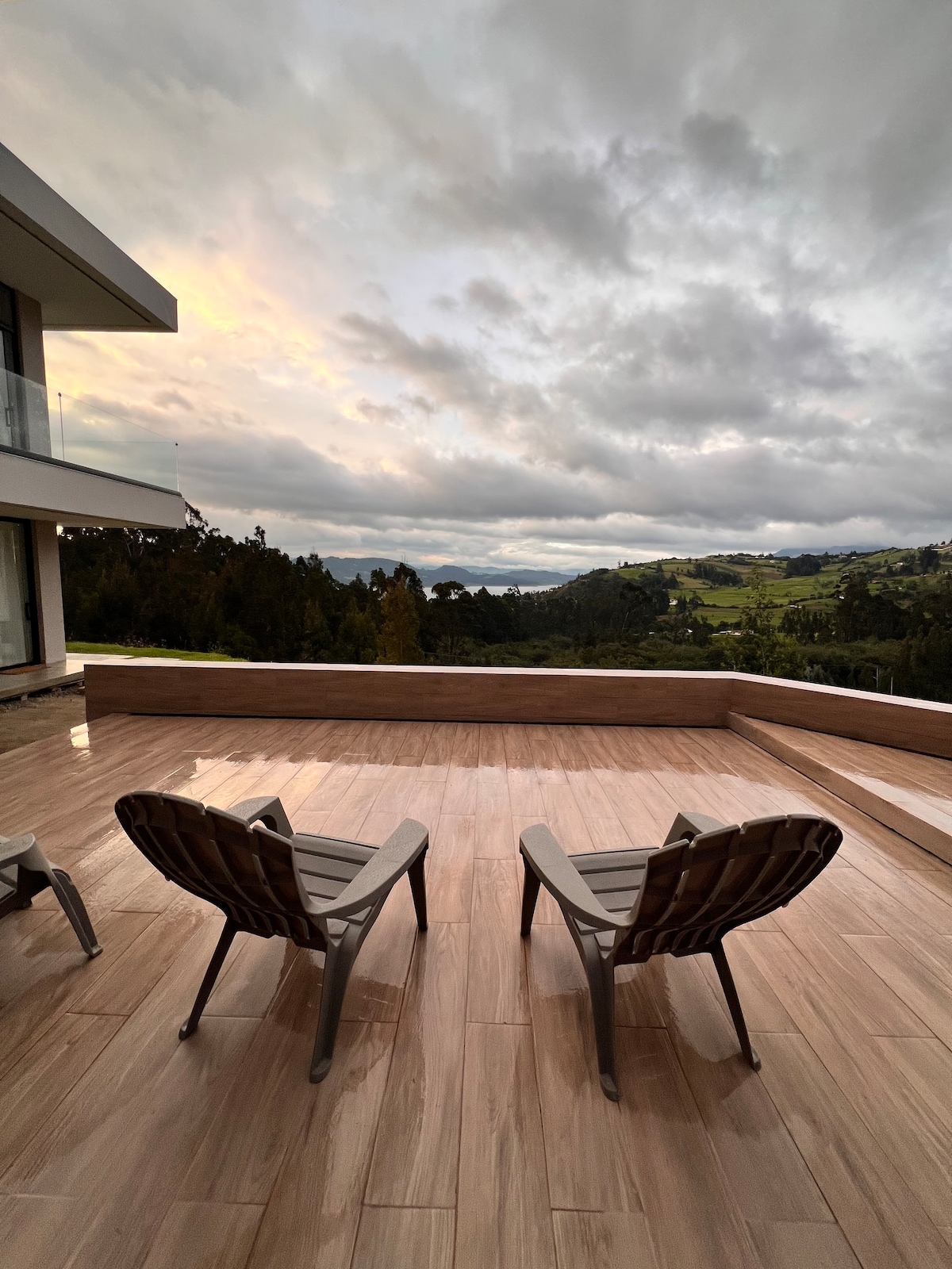 Two comfortable reclining chairs are positioned on a spacious wooden deck, overlooking a stunning landscape of rolling hills and valleys. The softly illuminated sky hints at evening, with clouds scattered above, creating a serene atmosphere.