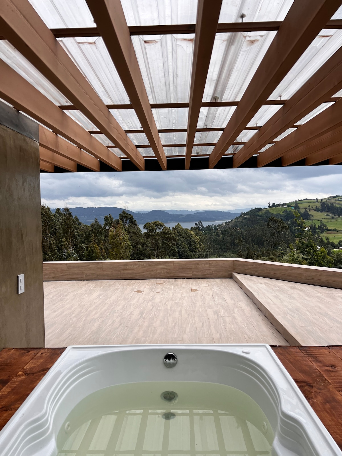 A private outdoor jacuzzi is positioned in the foreground, with a wooden deck and panoramic views of the surrounding mountains and lush greenery. A pergola covers part of the space, allowing natural light to filter through while providing an inviting atmosphere.