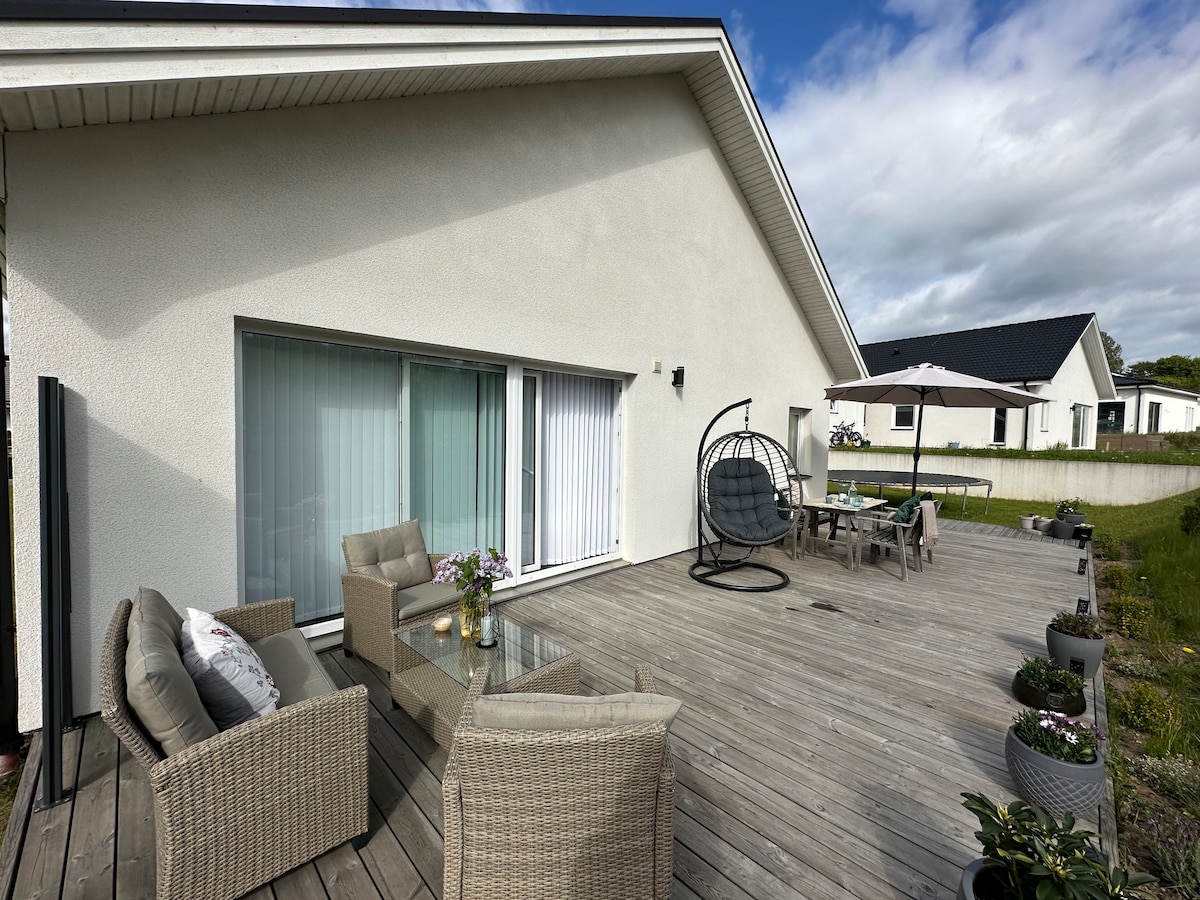 An outdoor terrace features a comfortable seating area with woven chairs and a glass table. A hanging chair adds a relaxed touch, while a dining table under an umbrella accommodates outdoor meals. Planters with greenery adorn the space, set against a well-kept wooden deck.