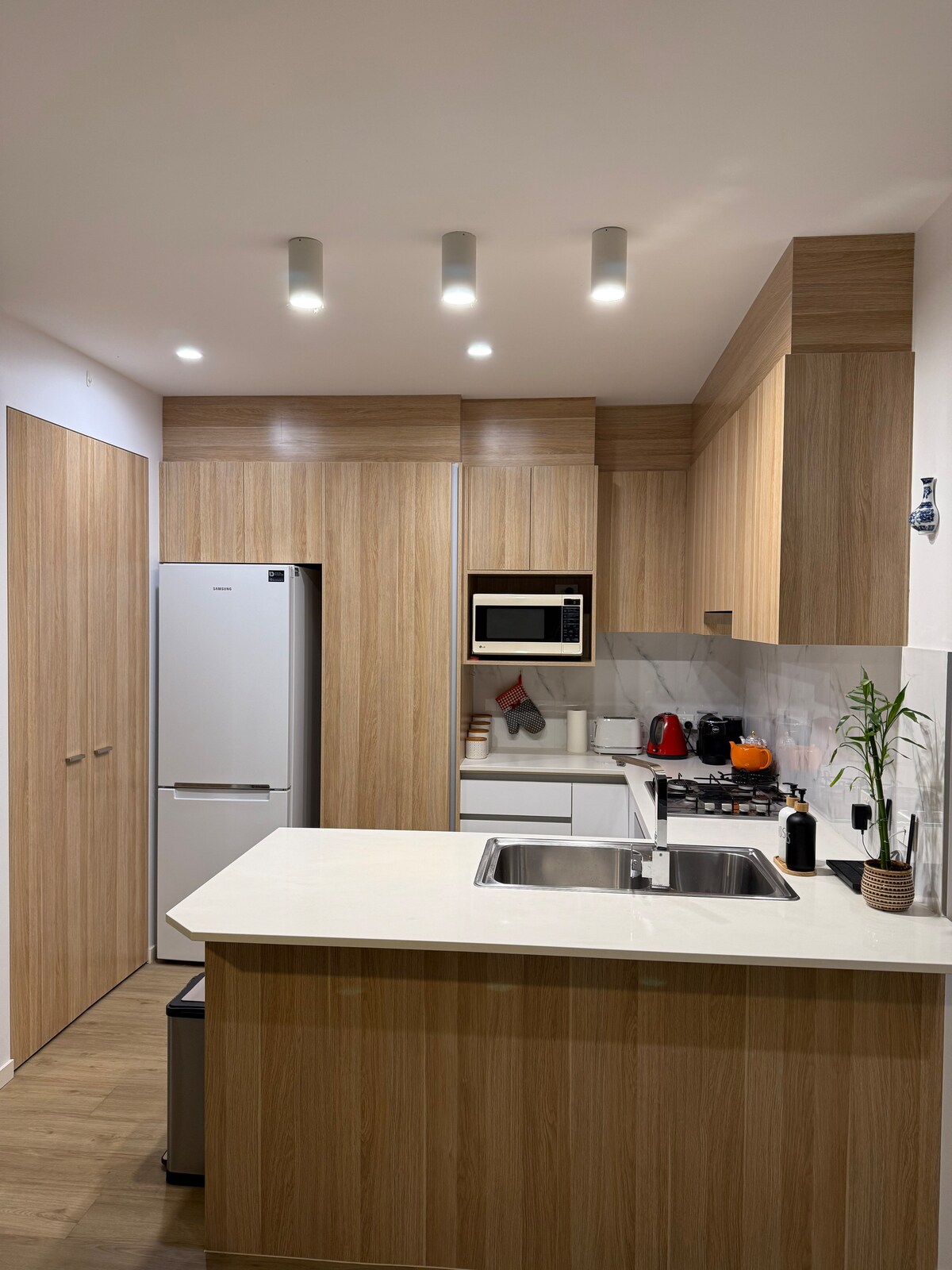 A modern kitchen is displayed with light wooden cabinetry and sleek finishes. Stainless steel appliances, including a refrigerator and microwave, are integrated. A gas stove is situated near a small sink, while ample counter space is available for preparation. Soft overhead lighting provides a bright atmosphere.