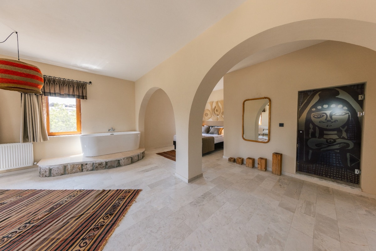 A spacious room is highlighted by arched doorways and a distinctive design. A large freestanding bathtub is positioned near a window, and a cozy seating area is visible in the background. A unique mirror and a patterned rug add character to the space.