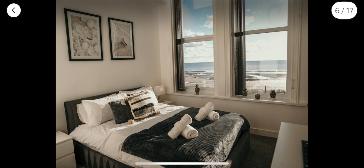 A serene bedroom is highlighted by expansive windows offering views of the sea. The spacious bed is adorned with decorative pillows and a plush blanket, while two rolled towels are neatly placed on top. Minimalist art frames and small plants accent the room.