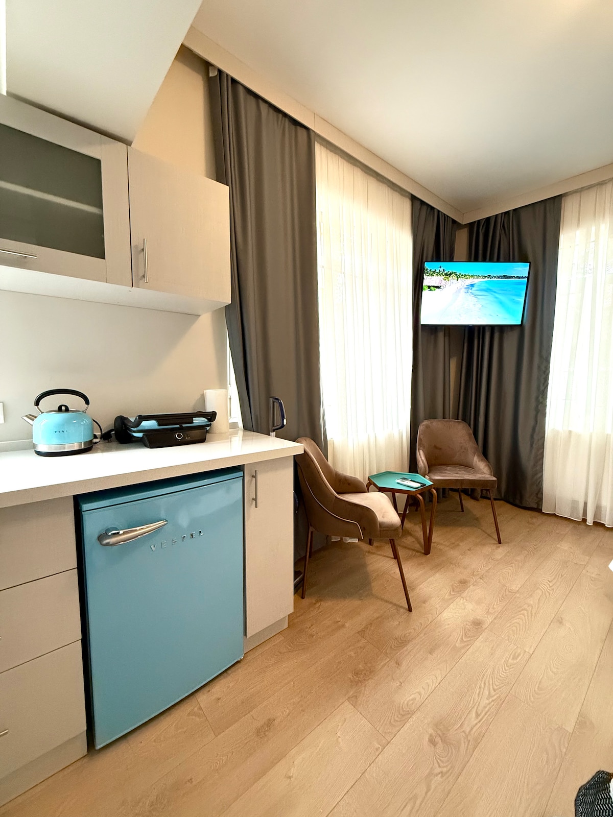 A cozy kitchen area features a blue refrigerator and a countertop with a kettle and cooking appliance. Two stylish chairs are positioned nearby, with a television mounted on the wall displaying a beach scene, complemented by bright curtains allowing natural light to enter.
