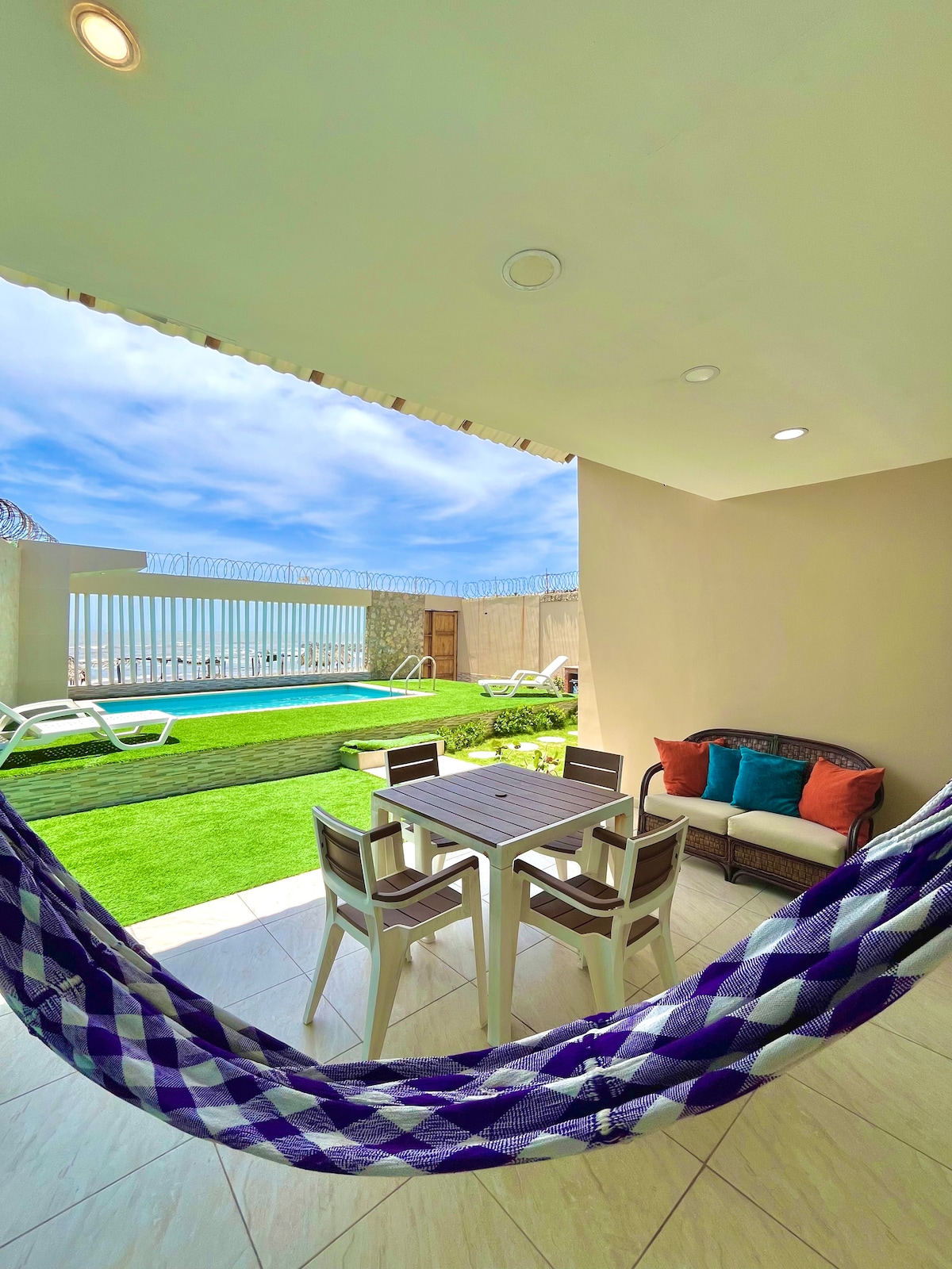 A shaded outdoor space is depicted, featuring a square dining table accompanied by four chairs. A cozy seating area with colorful cushions is visible. Beyond, a private pool glistens under a clear sky, framed by lush green grass and a safety railing.