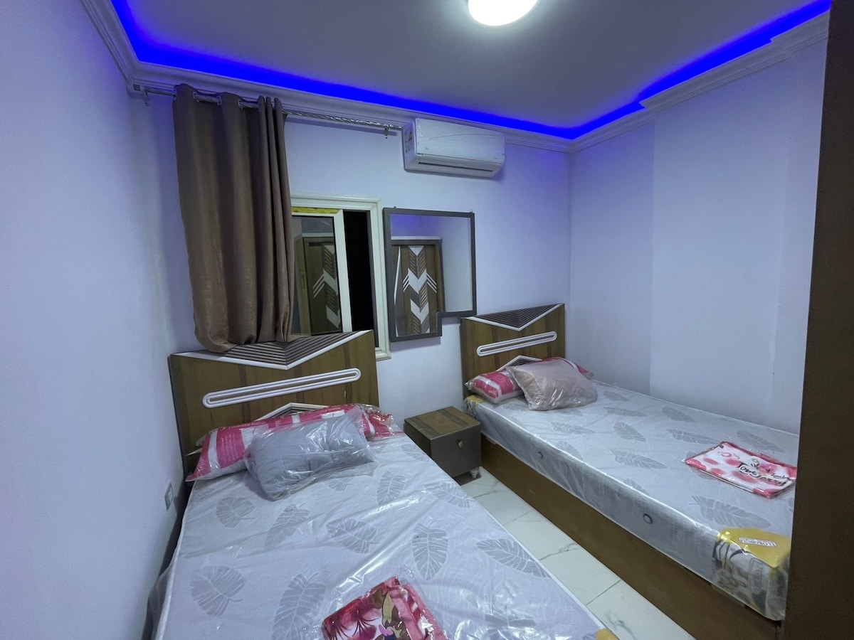 The room features two separate beds with modern headboards, positioned beside a small nightstand. A mirror is mounted on one wall, reflecting the space. Soft drapes frame a window, and ambient lighting is provided by a recessed ceiling fixture.